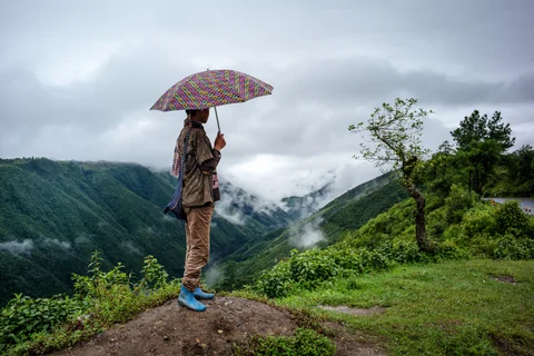 A man holding an umbrella waits for public transport by the side of a road in Cherrapunji. (Photographer: Sanjit Das/Bloomberg) 
