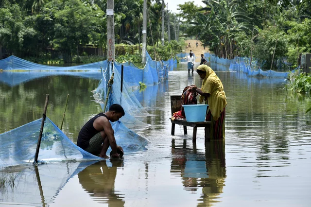 An Indian woman washes clothes next to man cleaning a pot along a flooded road following heavy monsoon rains at Kaljhar village in Barpeta district of Assam