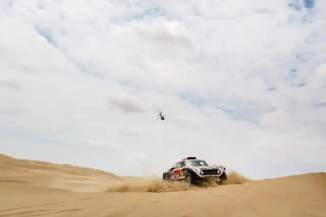 A Red Bull car struggles in the thick sand as a chopper flies by above.