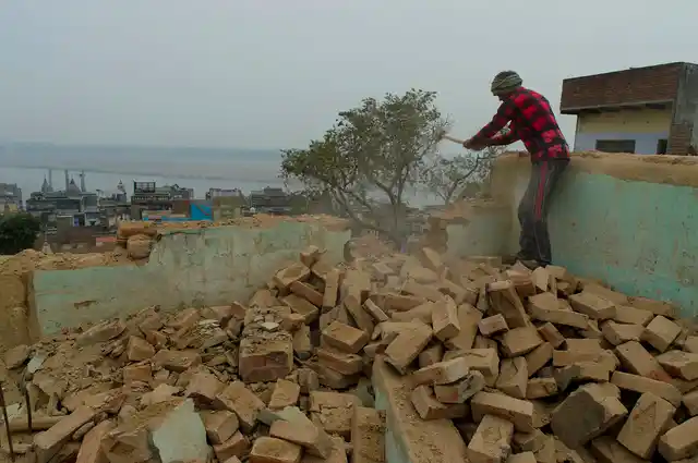 A worker breaks the parapet of a house. The Ganges is visible in the backdrop.