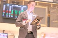 A stock trader in a jacket uses a tablet on the floor of the New York Stock Exchange, with digital stock tickers displaying red and green numbers in the background.