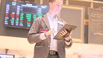 A stock trader in a jacket uses a tablet on the floor of the New York Stock Exchange, with digital stock tickers displaying red and green numbers in the background.