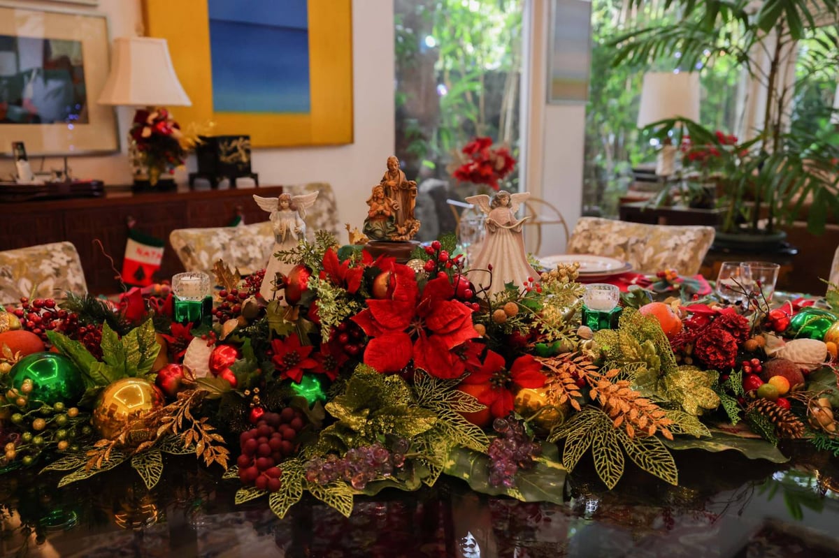 A centerpiece of poinsettias, tinsel balls , pine cones, berries and hollies adorns the square formal dining table