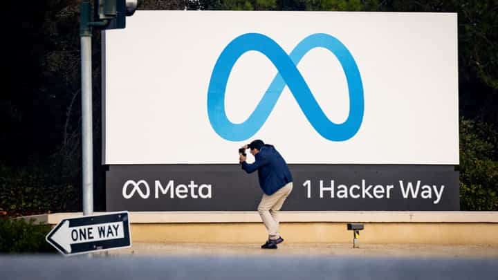A man takes a photo in front of the Meta headquarters sign at 1 Hacker Way in Menlo Park, California, featuring the Meta logo and company name.