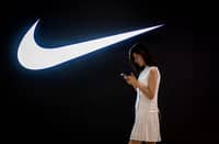 FILE PHOTO: A woman browses her phone while walking past a Nike logo inside a shopping mall. (Photo by NICOLAS ASFOURI / AFP)
