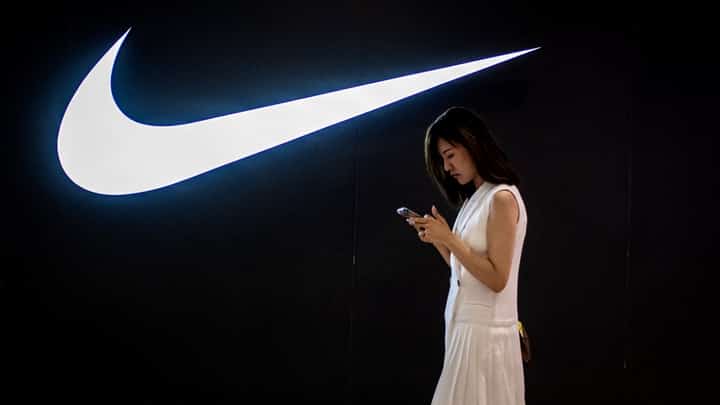 FILE PHOTO: A woman browses her phone while walking past a Nike logo inside a shopping mall. (Photo by NICOLAS ASFOURI / AFP)