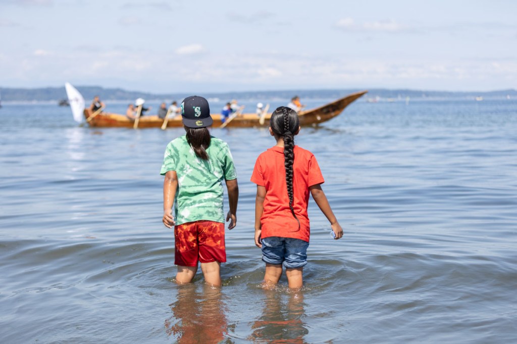 Two youth watch canoe families approach Alki Beach during Paddle to Muckleshoot.