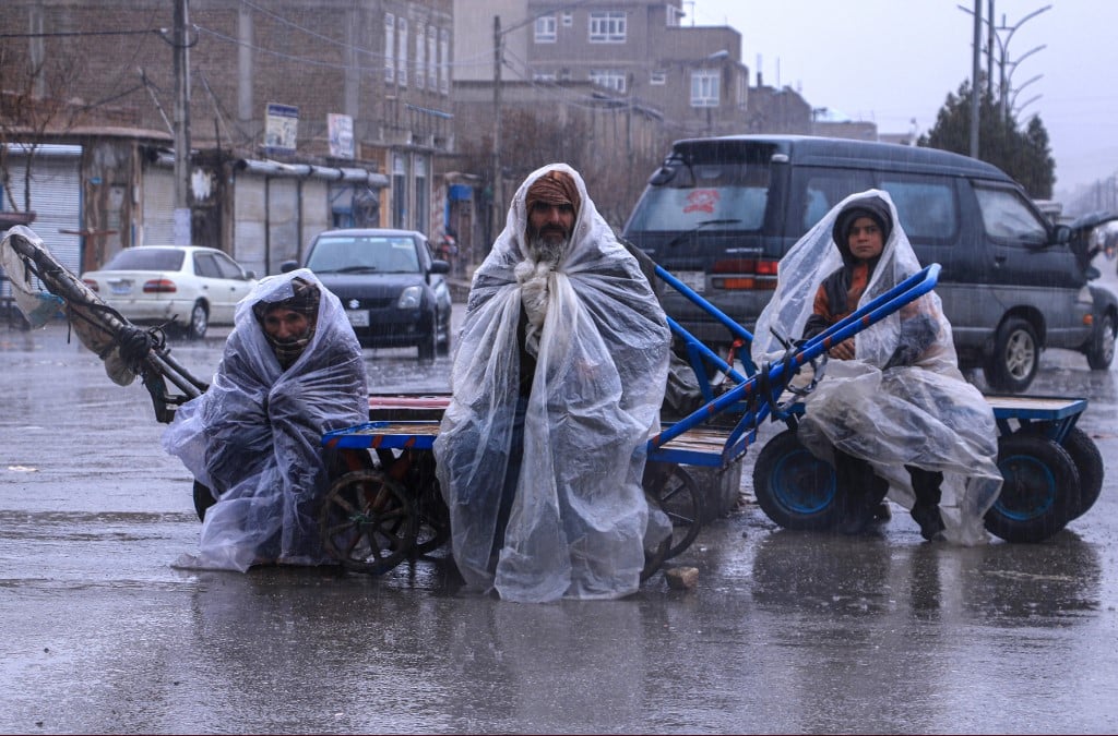 Afghan workers cover themselves with plastic sheets during the rainfall as they sit on their hand carts along a road in Herat on 12 March 2024.