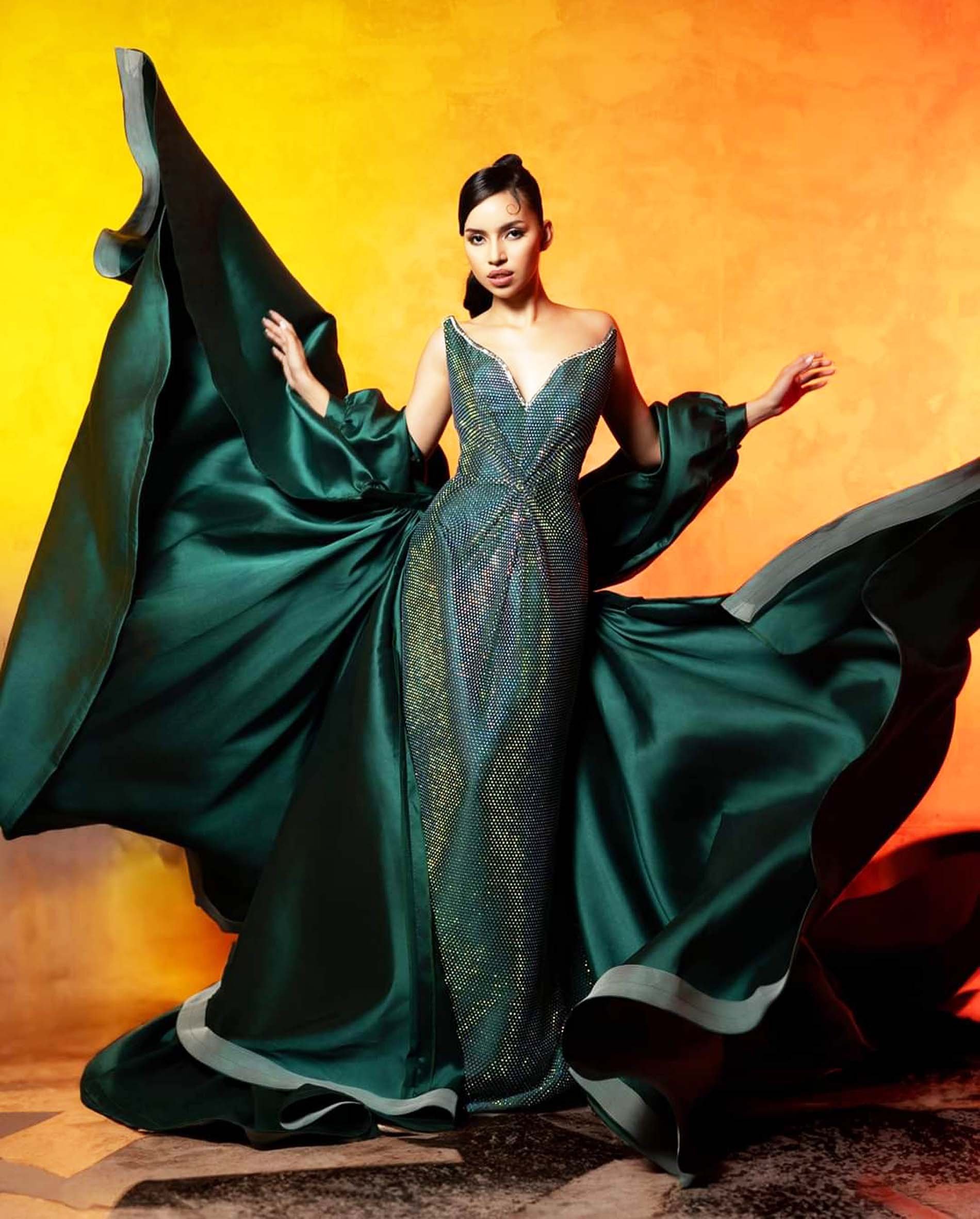 Model in a bejeweled emerald gown with cape.