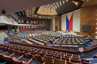 Wide view of the plenary hall of the Philippine House of Representatives at the Batasang Pambansa Complex, with empty seats and a large Philippine flag displayed at the front.