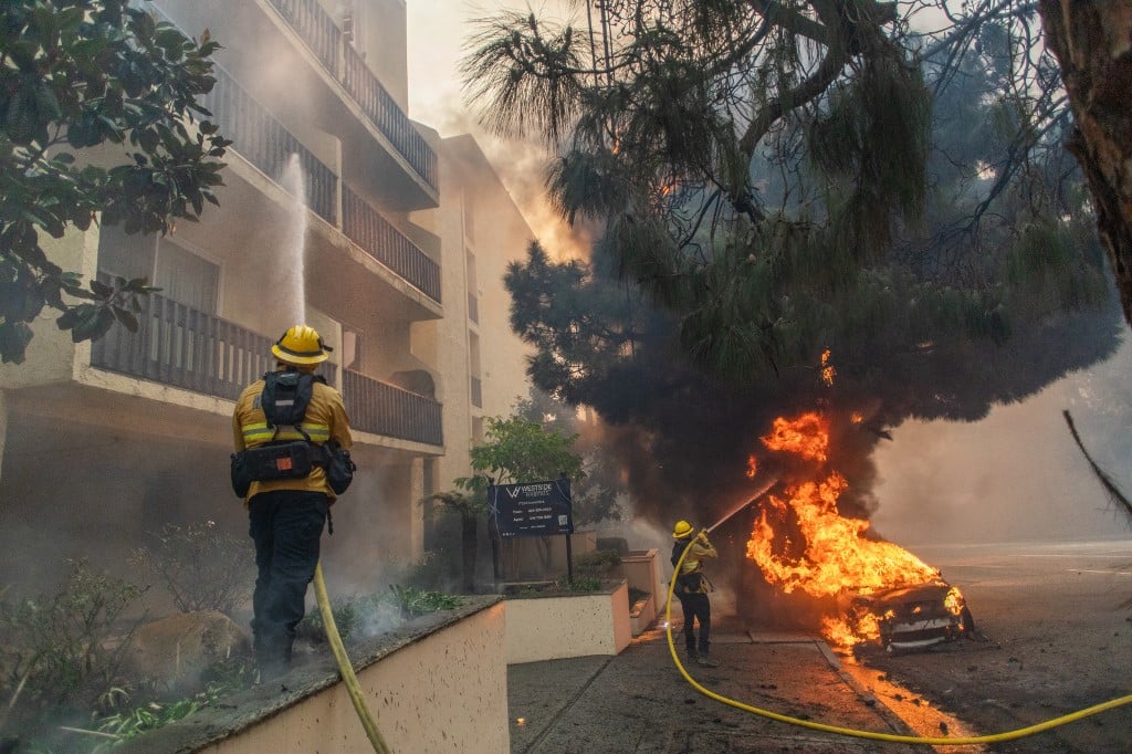 Firefighters sprays water towards a car and a complex building as flames from the Palisades Fire burn on Sunset Boulevard during a powerful windstorm on January 7, 2025 in Pacific Palisades, California. The fast-moving wildfire is threatening homes in the coastal neighborhood amid intense Santa Ana Winds and dry conditions in Southern California.