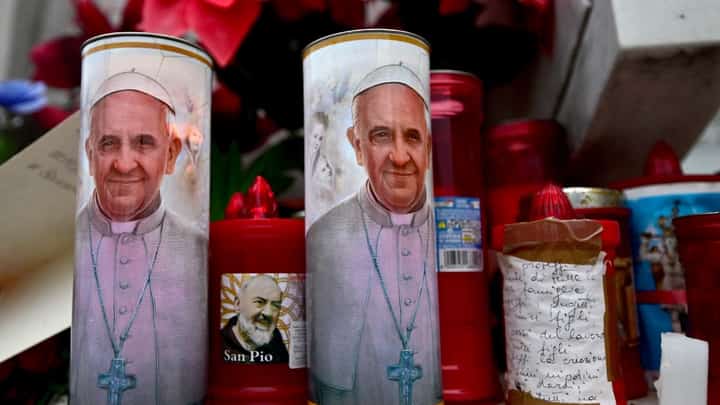 Candles with portraits of Pope Francis and Padre Pio are set at the bottom of a statue of Pope John Paul II outside the Gemelli Hospital where Pope Francis was hospitalized for tests and treatment for bronchitis in Rome, on February 19, 2025. Pope Francis, who has been diagnosed with pneumonia in both lungs, passed a "peaceful night", the Vatican said on 19 February 2025, amid growing concerns over the 88-year-old's condition.