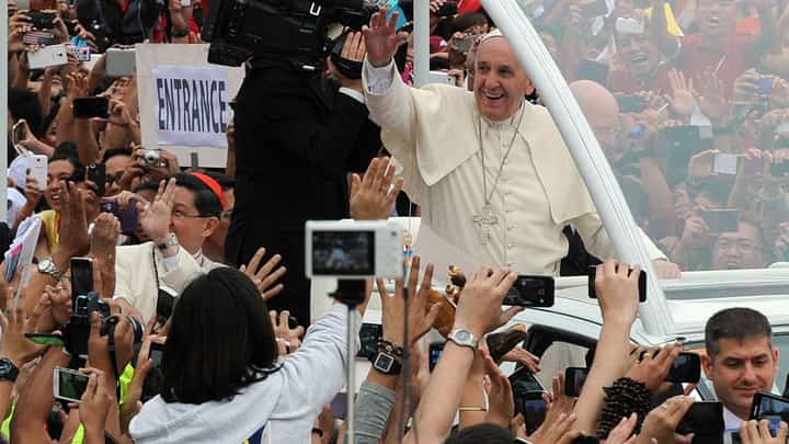 Pope Francis smiles and waves to a jubilant crowd of Filipino faithful during his visit to the Philippines, as hundreds raise their hands and phones to greet and capture the moment. Cardinal Luis Antonio Tagle is seen accompanying him in the popemobile.