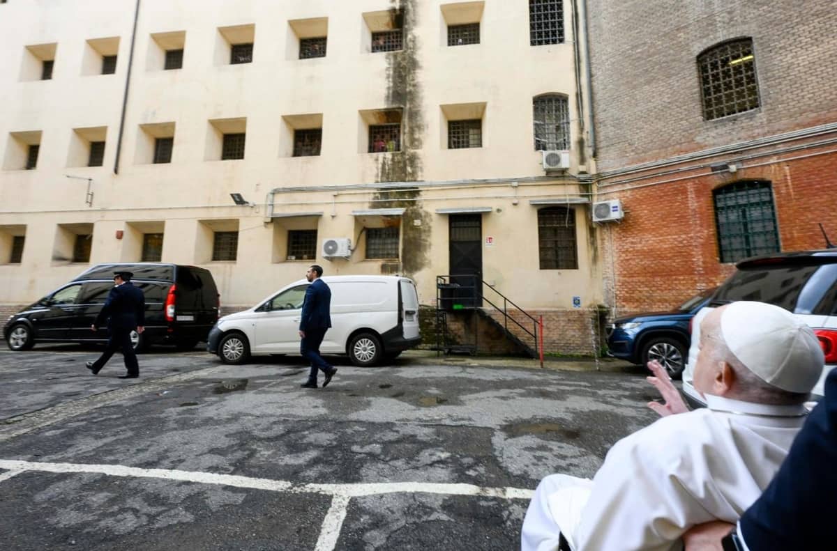 Pope Francis waves toward prison windows from his wheelchair in the courtyard of Regina Coeli Prison, as two guards walk ahead of him.