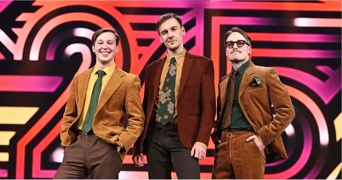 The Finnish comedy trio KAJ pose in retro-style brown and mustard-colored suits against a bold, colorful Eurovision 2025 stage backdrop. The three members smile confidently, exuding quirky charm ahead of their performance of Bara Bada Bastu.