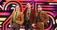The Finnish comedy trio KAJ pose in retro-style brown and mustard-colored suits against a bold, colorful Eurovision 2025 stage backdrop. The three members smile confidently, exuding quirky charm ahead of their performance of Bara Bada Bastu.