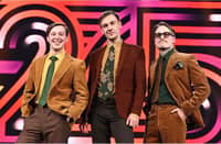 The Finnish comedy trio KAJ pose in retro-style brown and mustard-colored suits against a bold, colorful Eurovision 2025 stage backdrop. The three members smile confidently, exuding quirky charm ahead of their performance of Bara Bada Bastu.