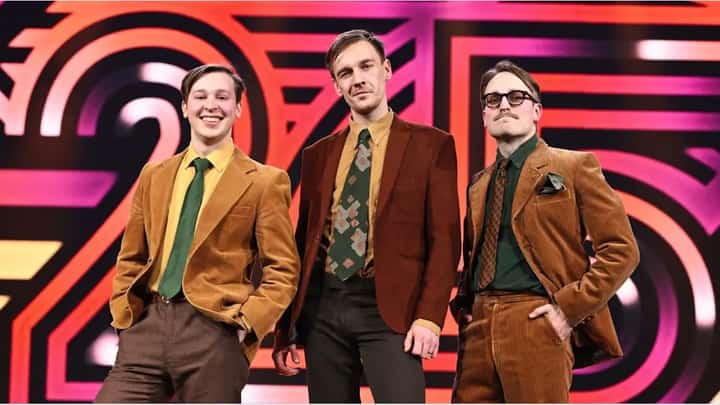 The Finnish comedy trio KAJ pose in retro-style brown and mustard-colored suits against a bold, colorful Eurovision 2025 stage backdrop. The three members smile confidently, exuding quirky charm ahead of their performance of Bara Bada Bastu.