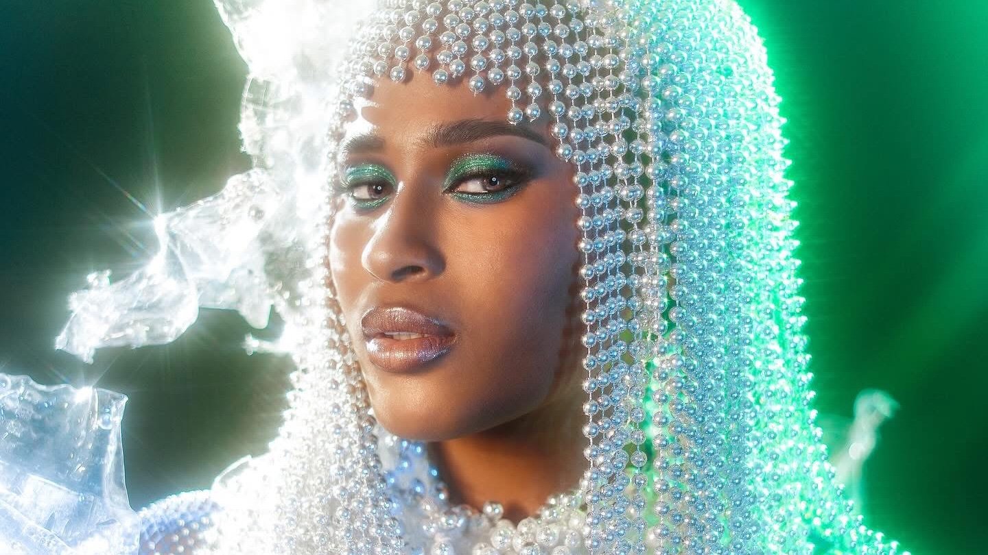 A close-up portrait of Alexie Brooks wearing a headpiece made of shimmering pearls, with green eyeshadow and glossy lips.