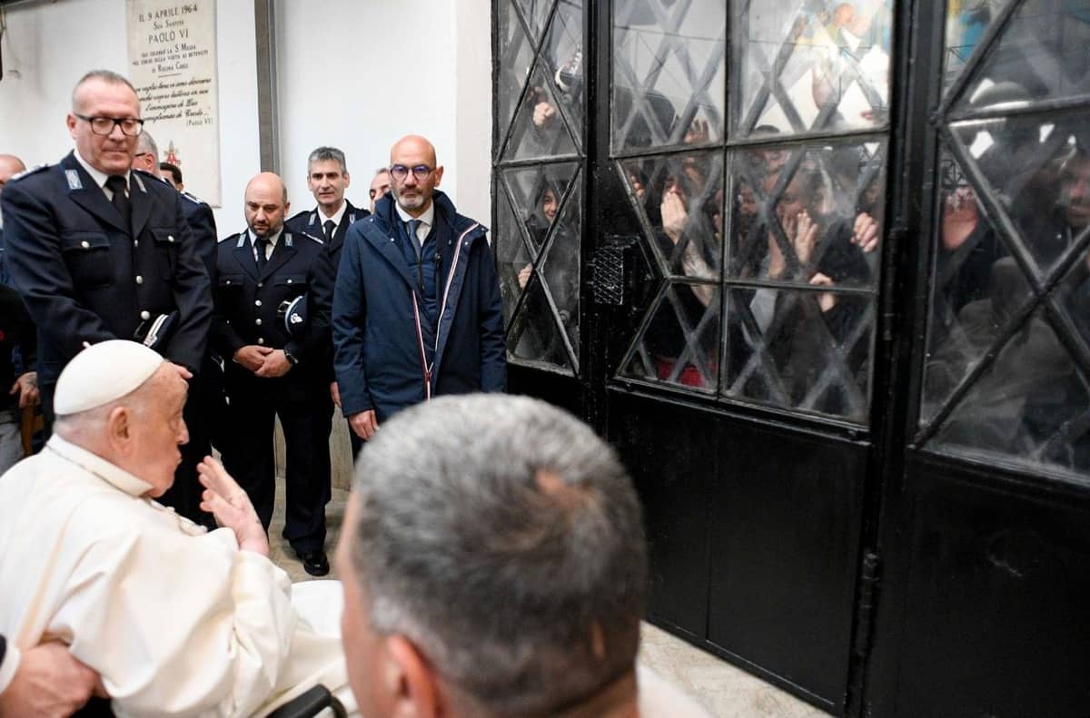 Pope Francis, seated in a wheelchair, gestures toward a group of inmates watching from behind a barred glass door inside Regina Coeli Prison, as prison guards stand nearby.