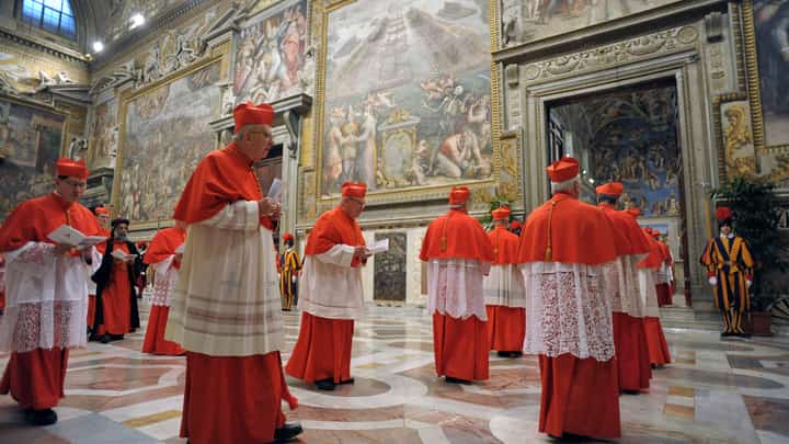Catholic cardinals dressed in red vestments walk solemnly through a fresco-lined corridor toward the Sistine Chapel as they prepare to enter the papal conclave.
