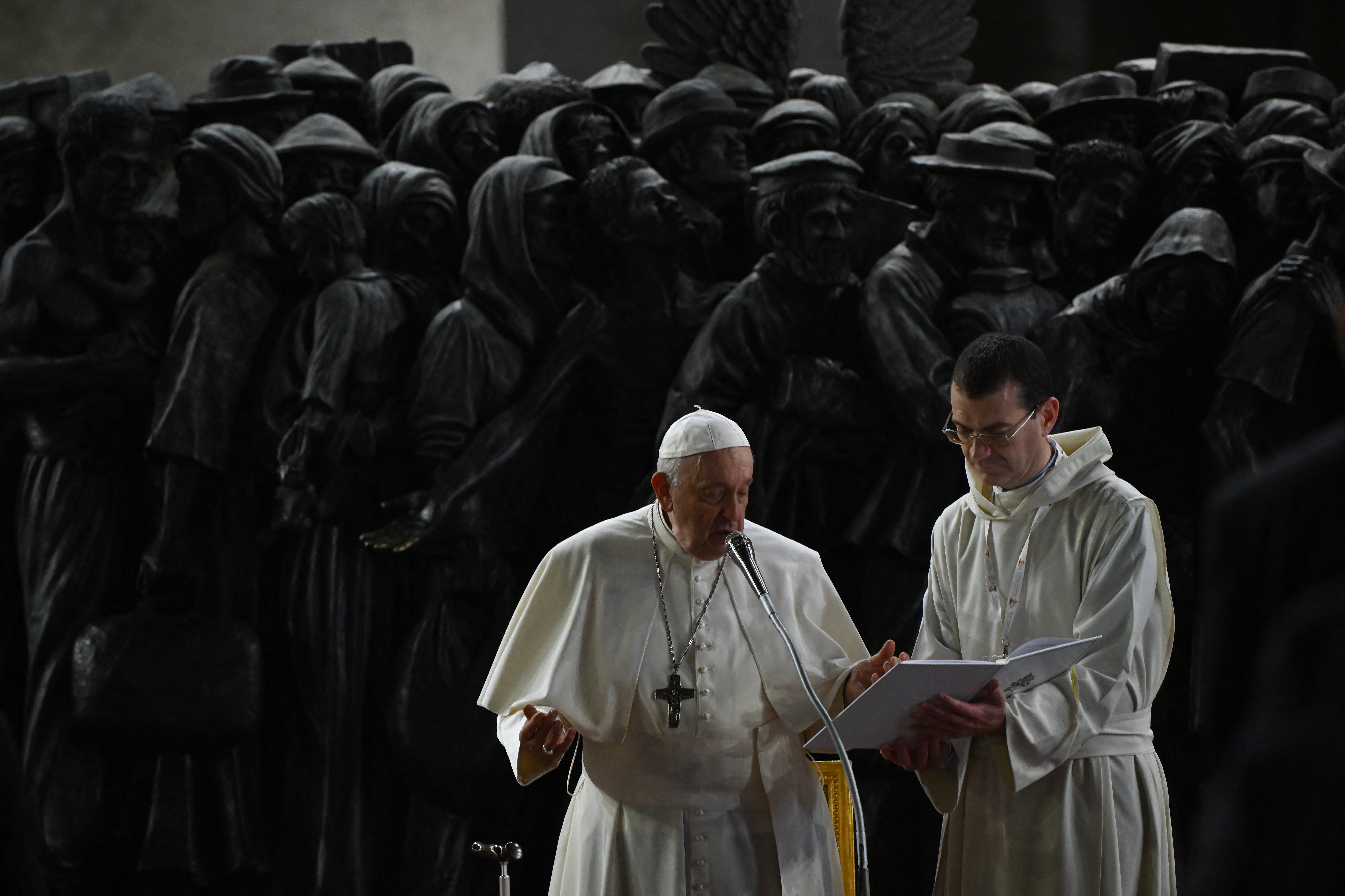 Pope Francis delivers a speech beside a cleric in front of a dark bronze sculpture depicting migrants and refugees, symbolizing solidarity and compassion.