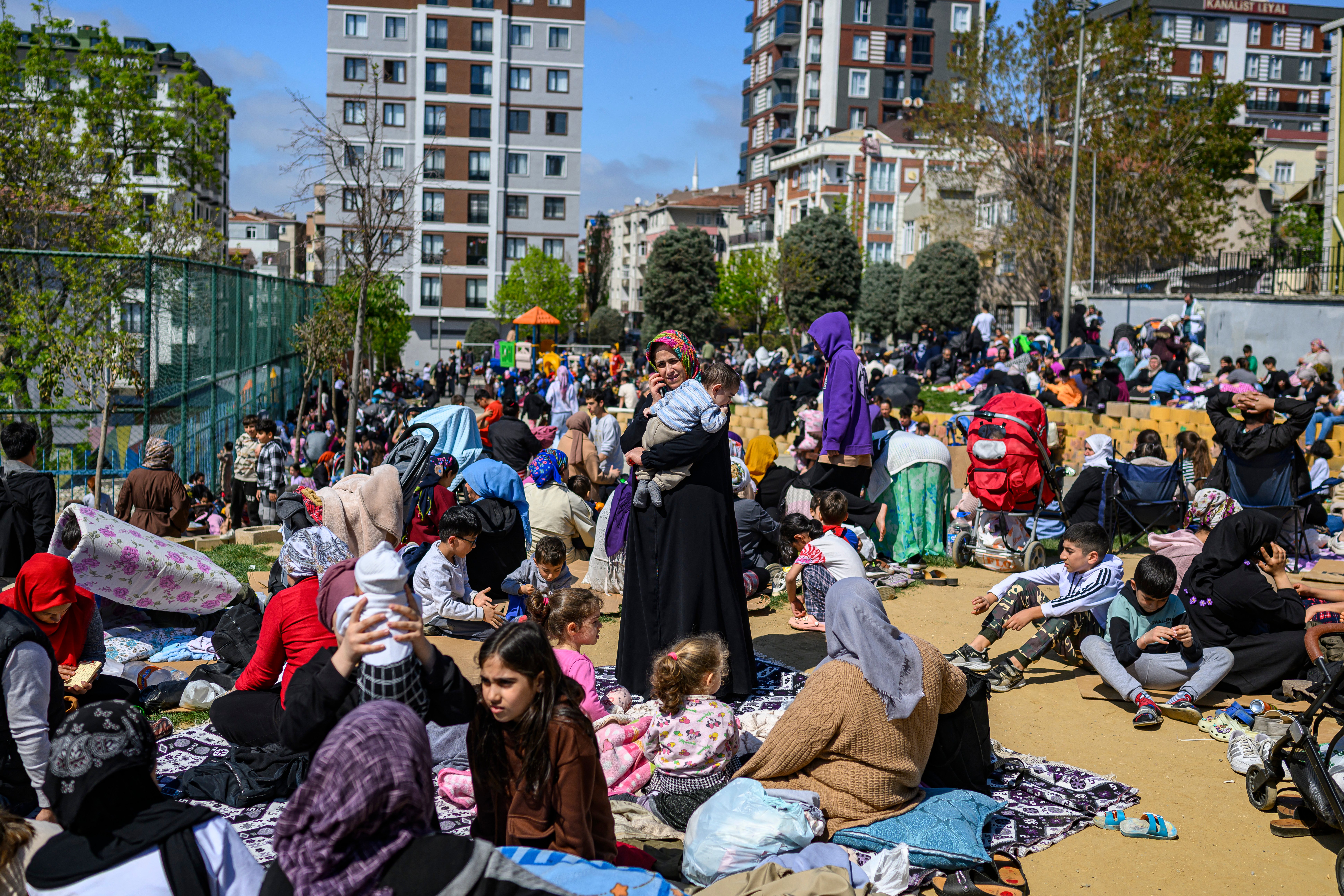 Crowds of Istanbul residents, including families with children, gather in a park during daylight after evacuating buildings due to a 6.2-magnitude earthquake, with people sitting on blankets and holding babies amid apartment buildings in the background.