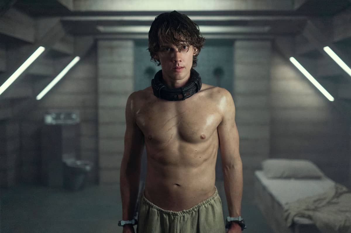Hunter Doohan as Tyler Galpin stands shirtless in a dim, futuristic holding cell, wearing a high-tech collar and wrist restraints in a scene from Netflix's "Wednesday" season 2.