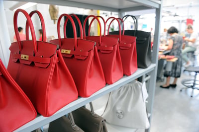 Rows of red Hermès Birkin bags are displayed on a shelf inside the Maroquinerie de la Tardoire, an Hermès workshop specializing in calfskin leather goods, in Montbron, southwestern France. Artisans can be seen working in the background.
