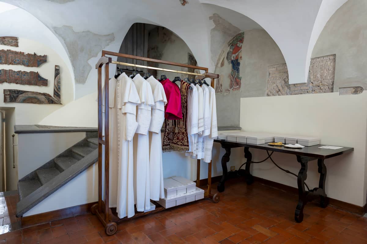 A rack of papal vestments, including white cassocks, a red mozzetta, and embroidered garments, stands in the Vatican's "Room of Tears" beside a wooden table lined with boxes, where the newly elected pope dresses for the first time after conclave.