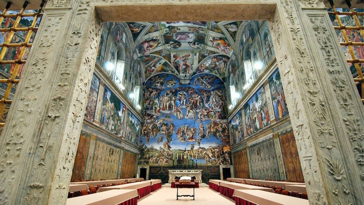 Interior view of the Sistine Chapel arranged for a papal conclave, with long tables covered in beige cloth and red skirting, set beneath Michelangelo’s iconic ceiling and “Last Judgment” fresco.