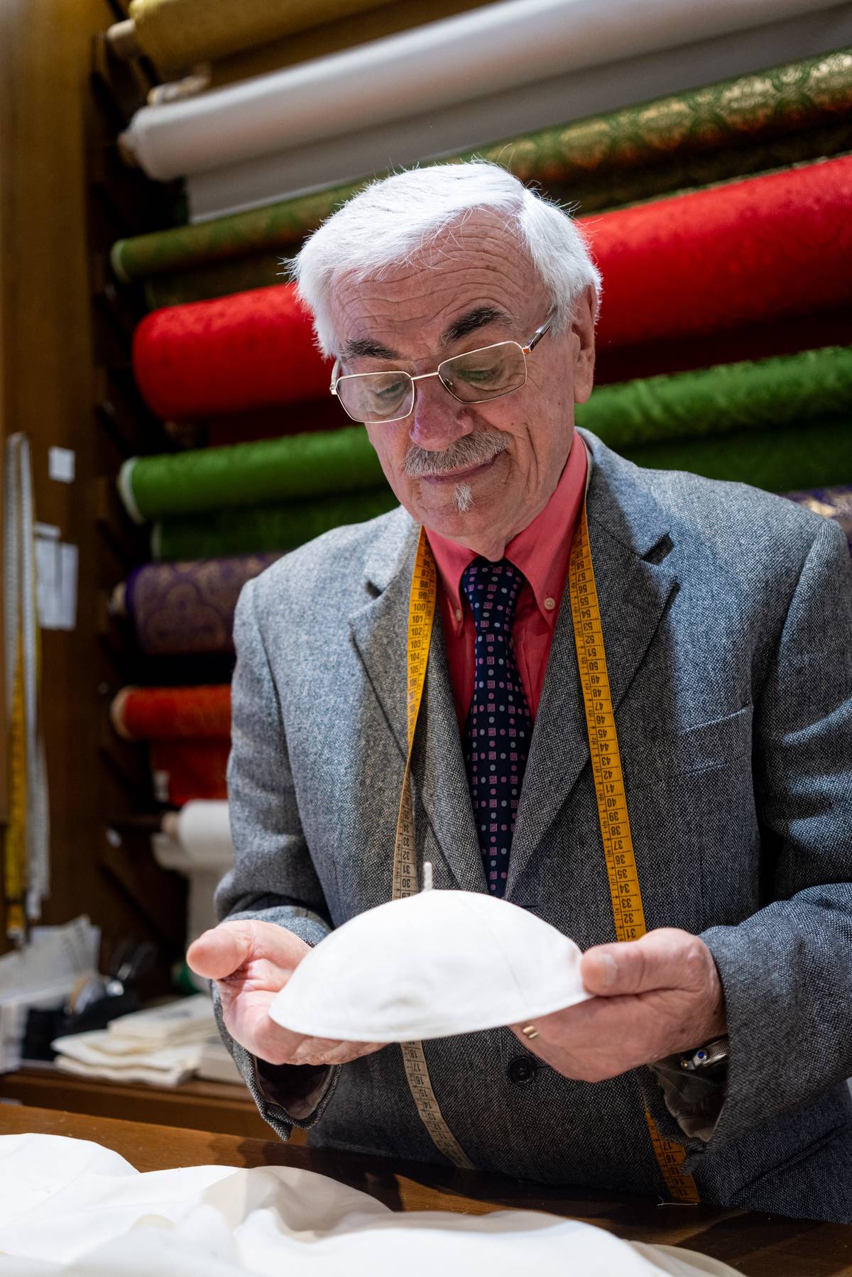 Italian tailor Raniero Mancinelli, 86, holds a white zucchetto (skullcap) he prepared for the future pope in his tailor shop in Rome, Italy, on 5 May 2025. Mancinelli has personally crafted cassocks for three popes. Rolls of liturgical fabric are stacked behind him.