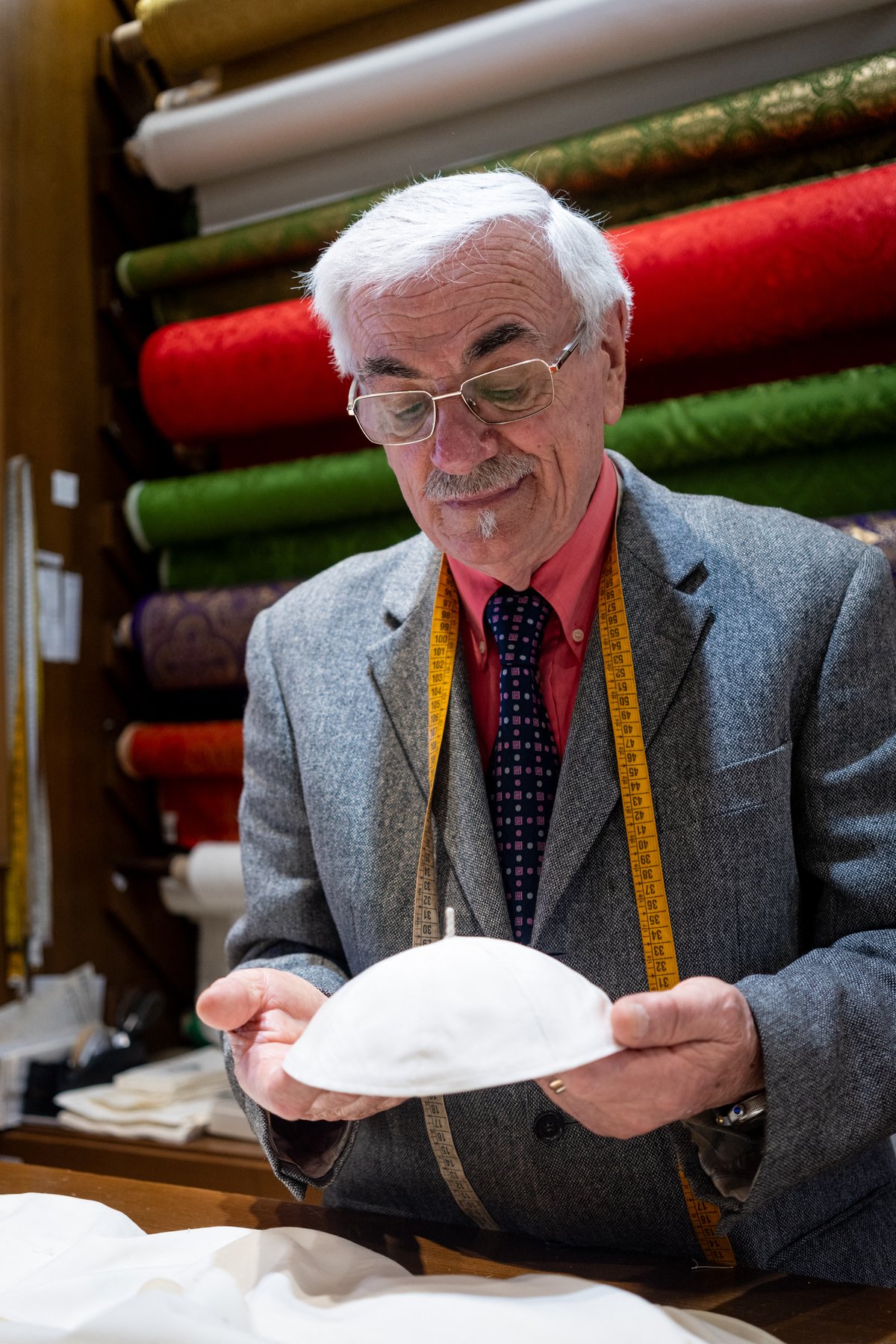 Italian tailor Raniero Mancinelli, 86, holds a white zucchetto (skullcap) he prepared for the future pope in his tailor shop in Rome, Italy, on 5 May 2025. Mancinelli has personally crafted cassocks for three popes. Rolls of liturgical fabric are stacked behind him.