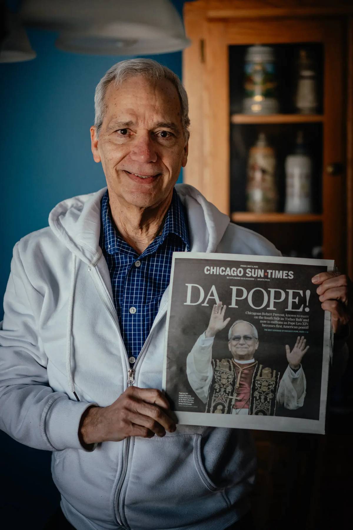 John Prevost, wearing a white hoodie and blue checkered shirt, smiles as he holds a Chicago Sun-Times newspaper with the headline “DA POPE!” featuring a photo of his brother, Pope Leo XIV, on the front page.