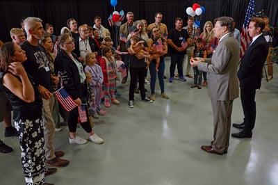 Deputy Secretary of State Christopher Landau and Homeland Security Deputy Secretary Troy Edgar address a group of Afrikaner refugees from South Africa during a welcome ceremony at Atlantic Aviation Dulles in Virginia on 12 May 2025. The group, including children, holds small U.S. flags as they listen.