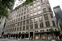 Exterior view of Saks Fifth Avenue in New York City, with American flags lining the historic building's facade. Shoppers are seen walking past display windows at street level.