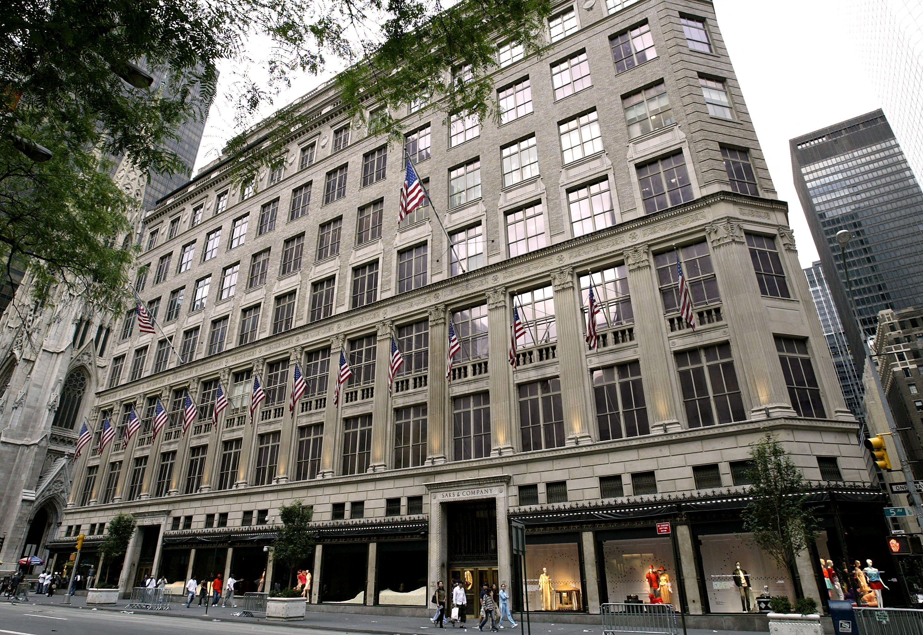Exterior view of Saks Fifth Avenue in New York City, with American flags lining the historic building's facade. Shoppers are seen walking past display windows at street level.
