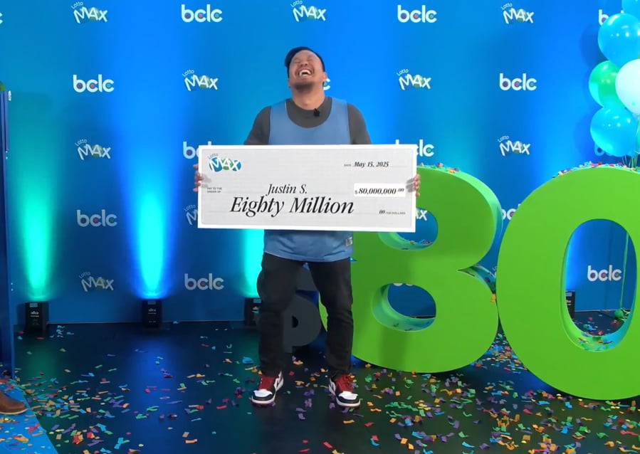 Justin Simporios from Surrey, B.C. laughs joyfully while holding a giant $80 million Lotto Max cheque during a celebration event hosted by BCLC. The moment marks the largest individual lottery win in Canadian history.