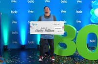 Justin Simporios from Surrey, B.C. laughs joyfully while holding a giant $80 million Lotto Max cheque during a celebration event hosted by BCLC. The moment marks the largest individual lottery win in Canadian history.