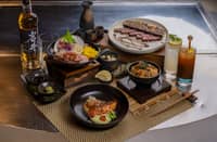 A luxurious five-course Japanese dinner set from Yakumi, featuring assorted sashimi, grilled Wagyu steak, miso-baked scallop, rice bowl, fresh edamame, dipping sauces, Japanese whisky, and two signature cocktails, beautifully plated on a teppanyaki-style table.