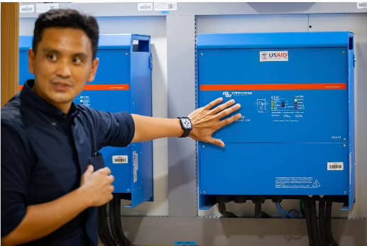 A USAID Energy Secure Philippines representative gestures toward a blue grid-forming inverter, explaining its functions. The device is part of a U.S.-donated mobile energy system designed to support electricity access in remote areas.