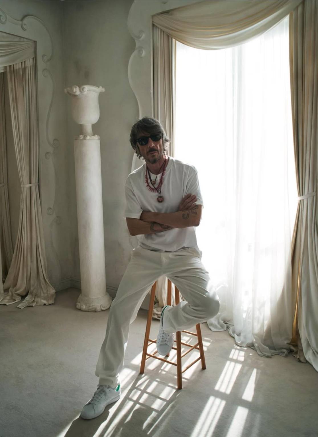Fashion designer Pierpaolo Piccioli sits on a wooden stool in a softly lit room with draped curtains, wearing sunglasses, white clothing, and layered necklaces. The portrait, taken by David Sims, was reposted by Balenciaga and Piccioli on Instagram after his appointment as the brand’s new creative director.