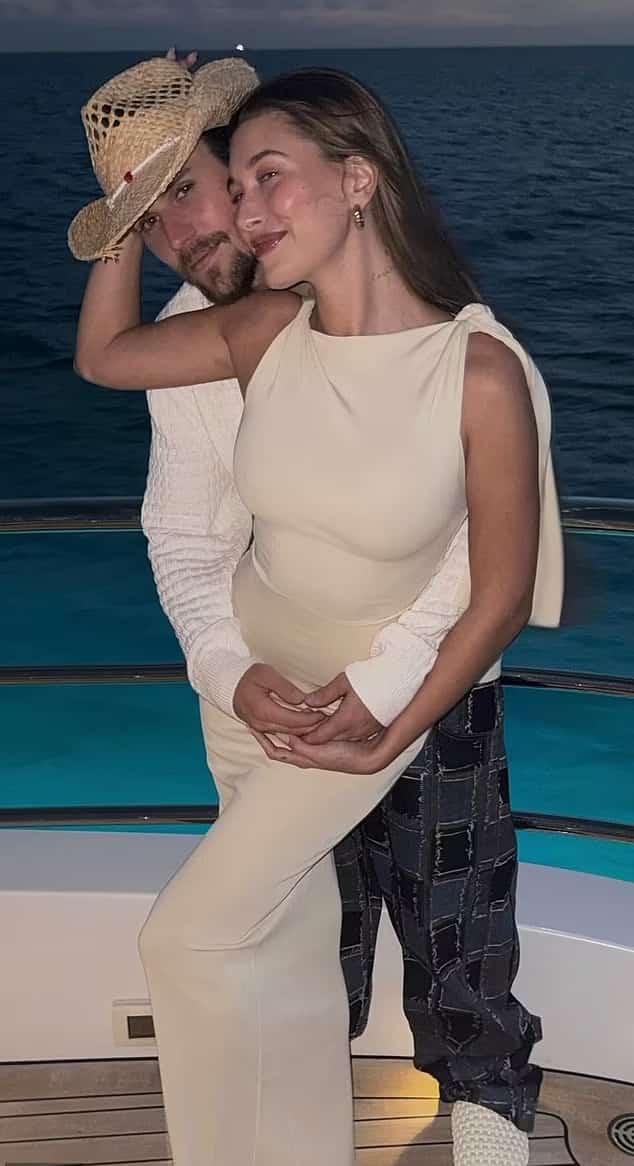Justin and Hailey Bieber share a tender moment on a yacht at sunset, with Justin wearing a straw hat and holding Hailey close from behind. Hailey leans back smiling, dressed in an elegant ivory outfit, with the ocean in the background.