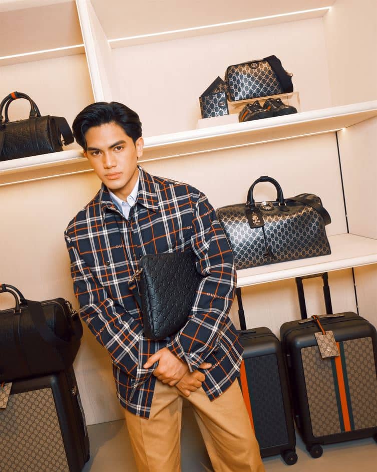Jarren Garcia dressed in a Gucci Cruise 2026 ensemble, featuring a blue-and-white tartan cotton padded overshirt, an azure striped cotton popeline shirt with fil coupé detailing, bisque double-cotton twill chino pants with web accents, and a medium GG Emblem accessory.