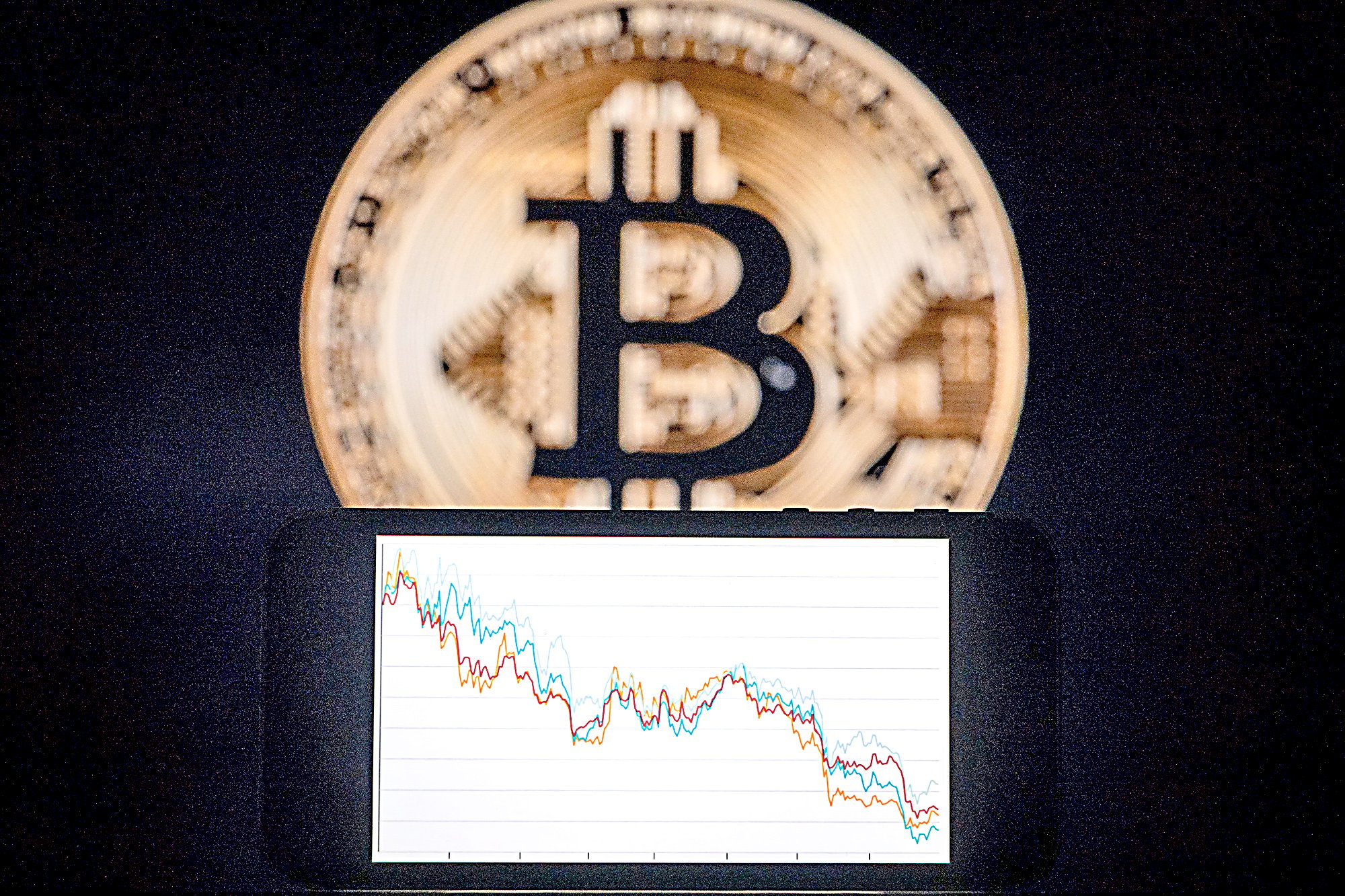 A PHONE with a downward trend in the stock market in front of a screen displaying a Bitcoin logo as the cryptocurrency’s value plummets.