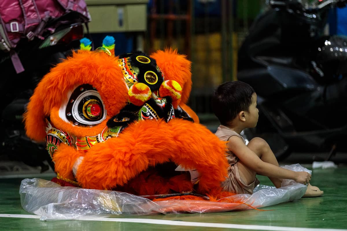 Jhayvee Sicat’s son sits beside a lion head costume during a practice session in downtown Manila on Friday, 6 February 2026. Sicat is a second-generation performer, who grew up in a family of dragon dance artists. He says he plans to pass his skills on to his children.