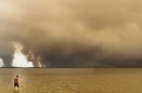 A BEACHGOER walks in the lake Lacanau from the beach of Moutchic in Lacanau, southwestern France on 24 July 2026, as clouds of smoke rise into the sky from a wildfire.