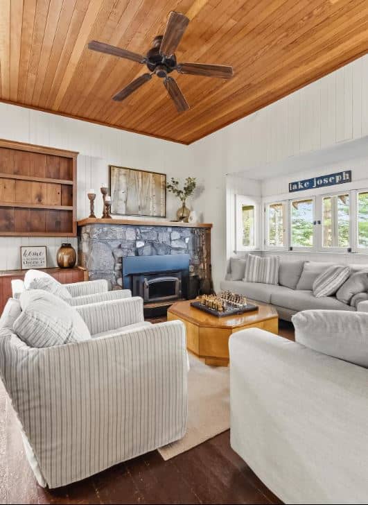 A stone fireplace anchors the living room with striped armchairs and a gray sofa beneath a wood-paneled ceiling fan.