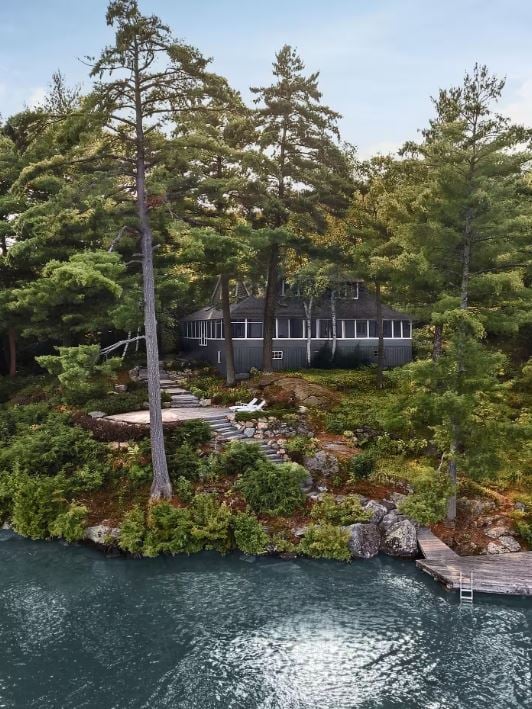 LAKESIDE view of a dark gray cottage framed by pine trees, stone steps, and a wooden dock on rocky shoreline.