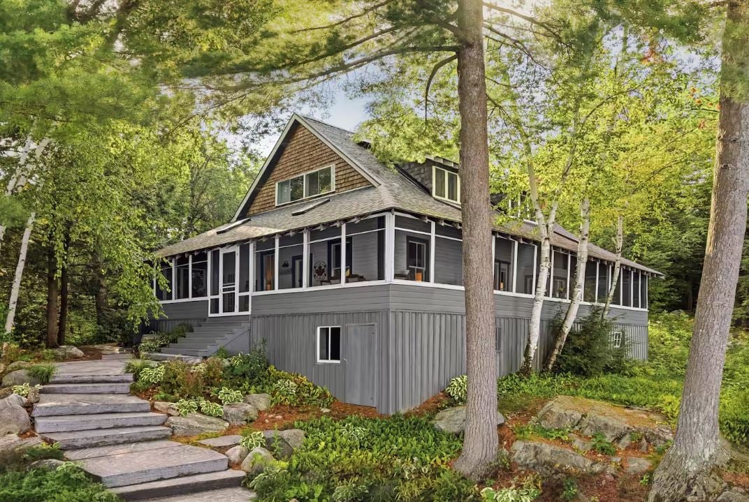 GRAY, wood-paneled cottage raised on stone steps, nestled among lush greenery and framed by natural rock landscaping.